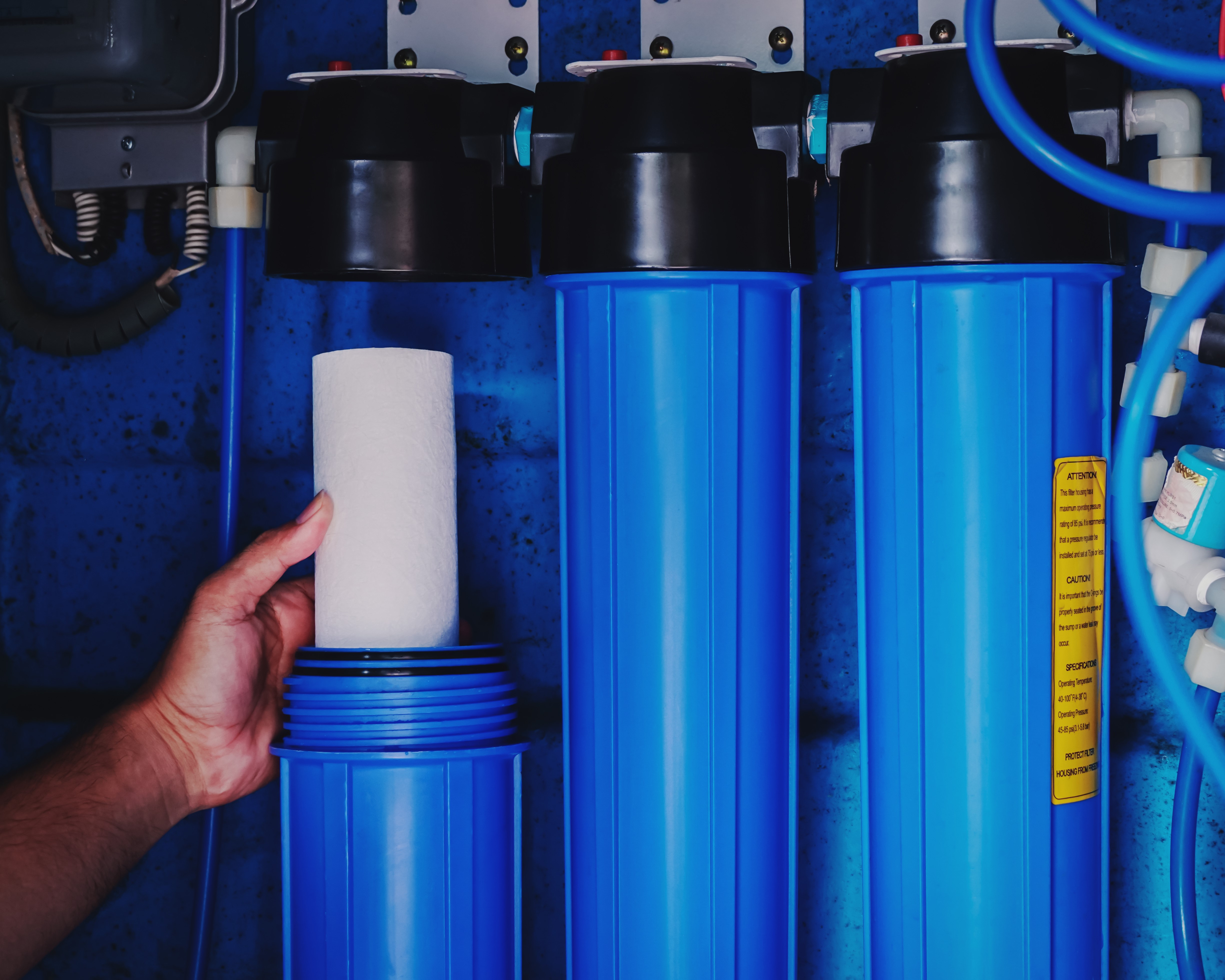 A person stands holding a water filter, positioned in front of blue water pipes, emphasising water purification efforts.